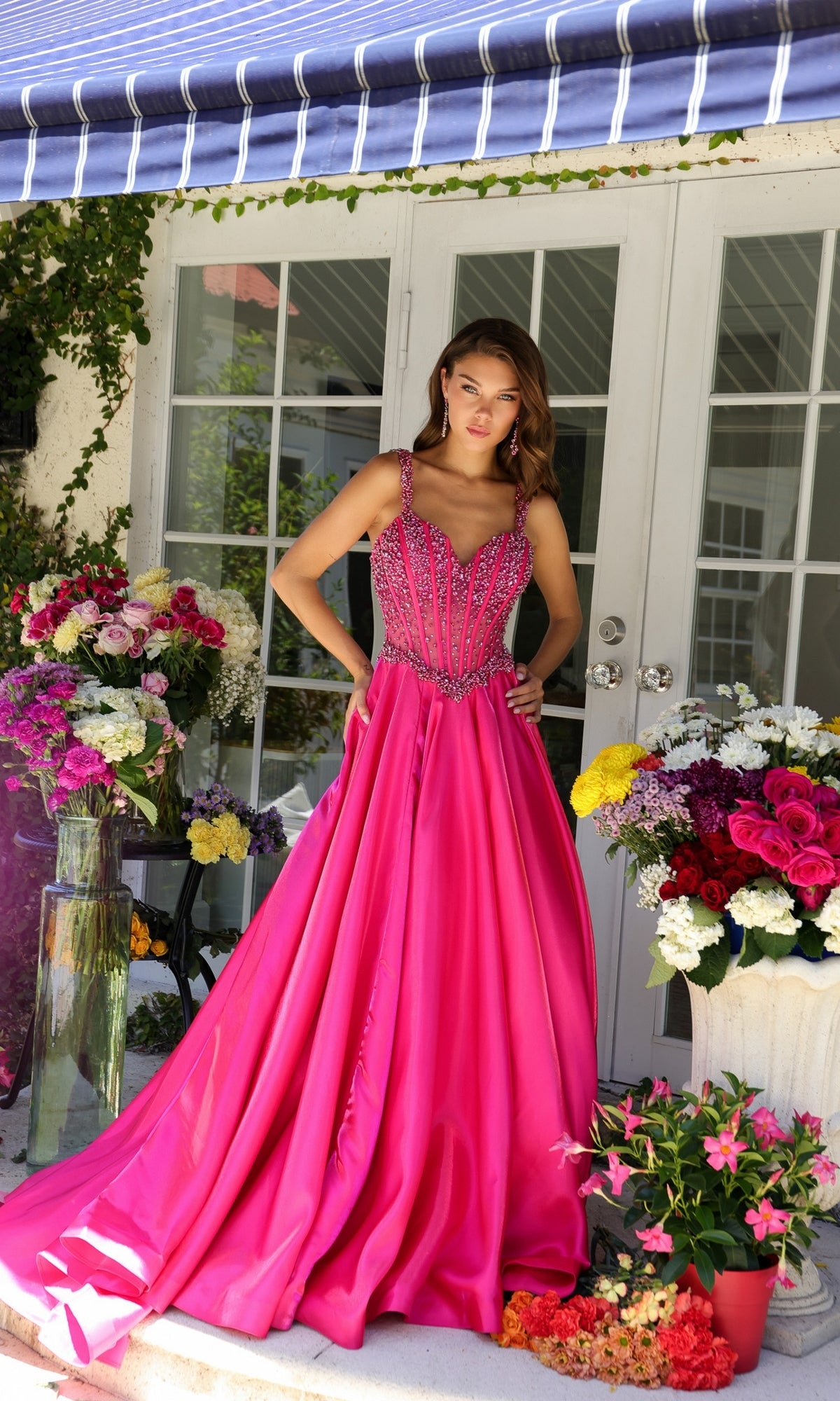 A woman wearing the Long Formal Dress 29521 by Ava Presley, a bright pink floor-length gown with a beaded corset bodice, stands on a flower-filled porch in front of white double doors beneath a blue-striped awning.